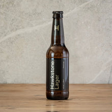 Load image into Gallery viewer, A single brown glass beer bottle labeled Hawkstone Lager stands upright on a wooden surface against a light stone wall.