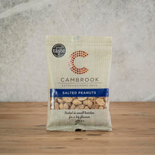 Load image into Gallery viewer, A bag of Cambrook Extraordinary Nuts Salted Peanuts standing upright on a wooden surface against a textured light gray wall.