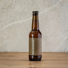 Load image into Gallery viewer, A single amber glass beer bottle with a brown paper label reading Hawkstone IPA, standing on a wooden surface against a light stone wall.
