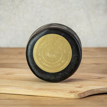 Load image into Gallery viewer, A round of Black Cow cheese with a gold label reading on a wooden cutting board against a textured light gray background.
