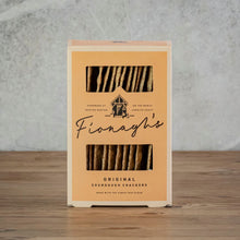 Load image into Gallery viewer, A boxed pack of Fionagh's Original Sourdough Crackers standing upright on a wooden surface against a textured light gray wall. 
