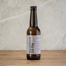 Load image into Gallery viewer, A single amber glass beer bottle with a white label reading Hawkstone 3.8 Pils, standing on a wooden surface against a textured light gray wall.

