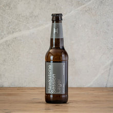 Load image into Gallery viewer, A bottle of Hawkstone Session Lager beer in an amber glass bottle with a gray label and dark cap, standing on a wooden table.
