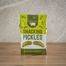 Load image into Gallery viewer, A pouch of Serious Pig Snacking Pickles sits on a wooden tabletop against a textured gray wall. 
